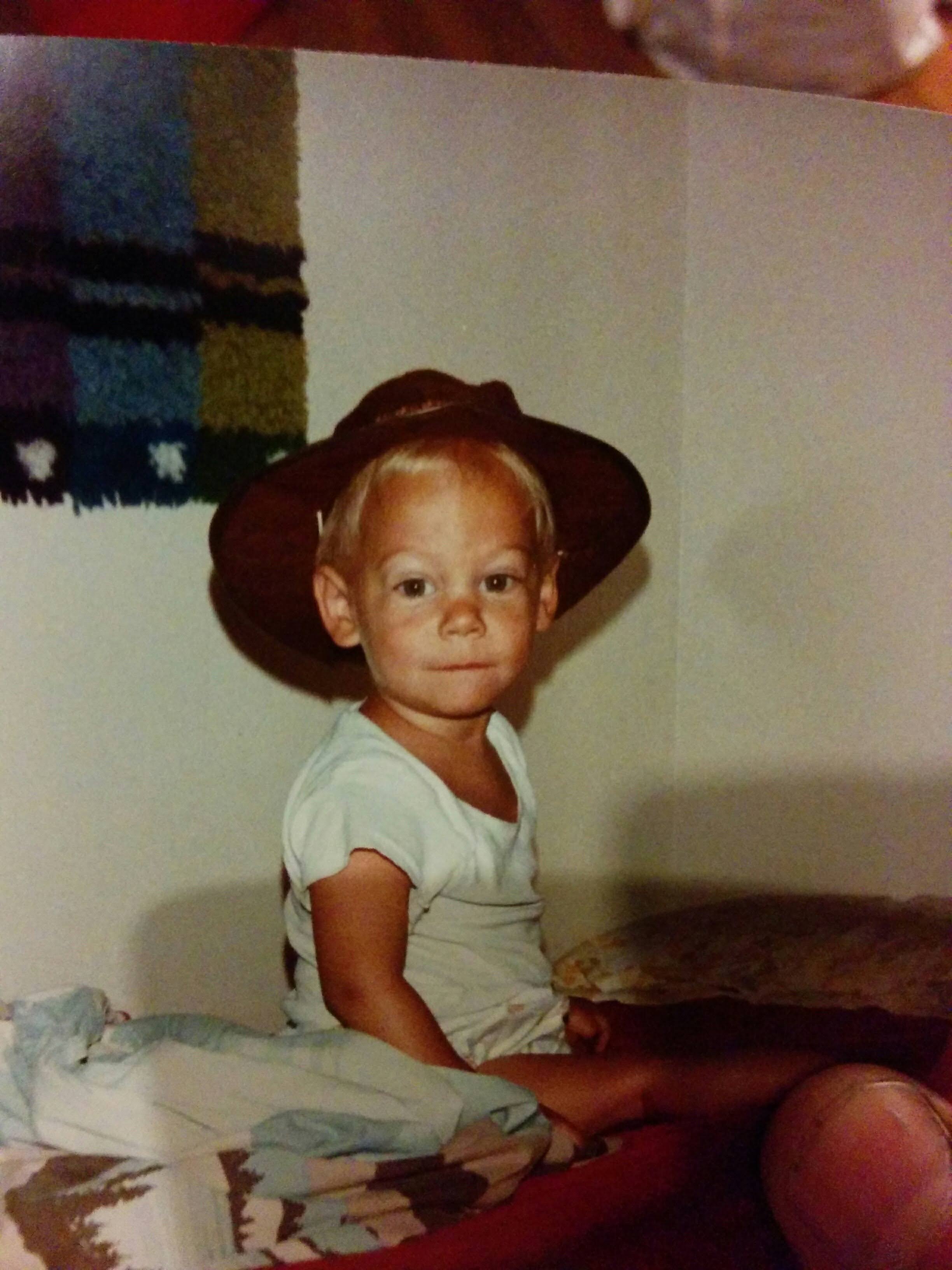 Levi at 2.5 years old wearing a cowboy hat and sitting on a bed. In the bottom right corner of the image you can see Levi's adult thumb holding the photograph.